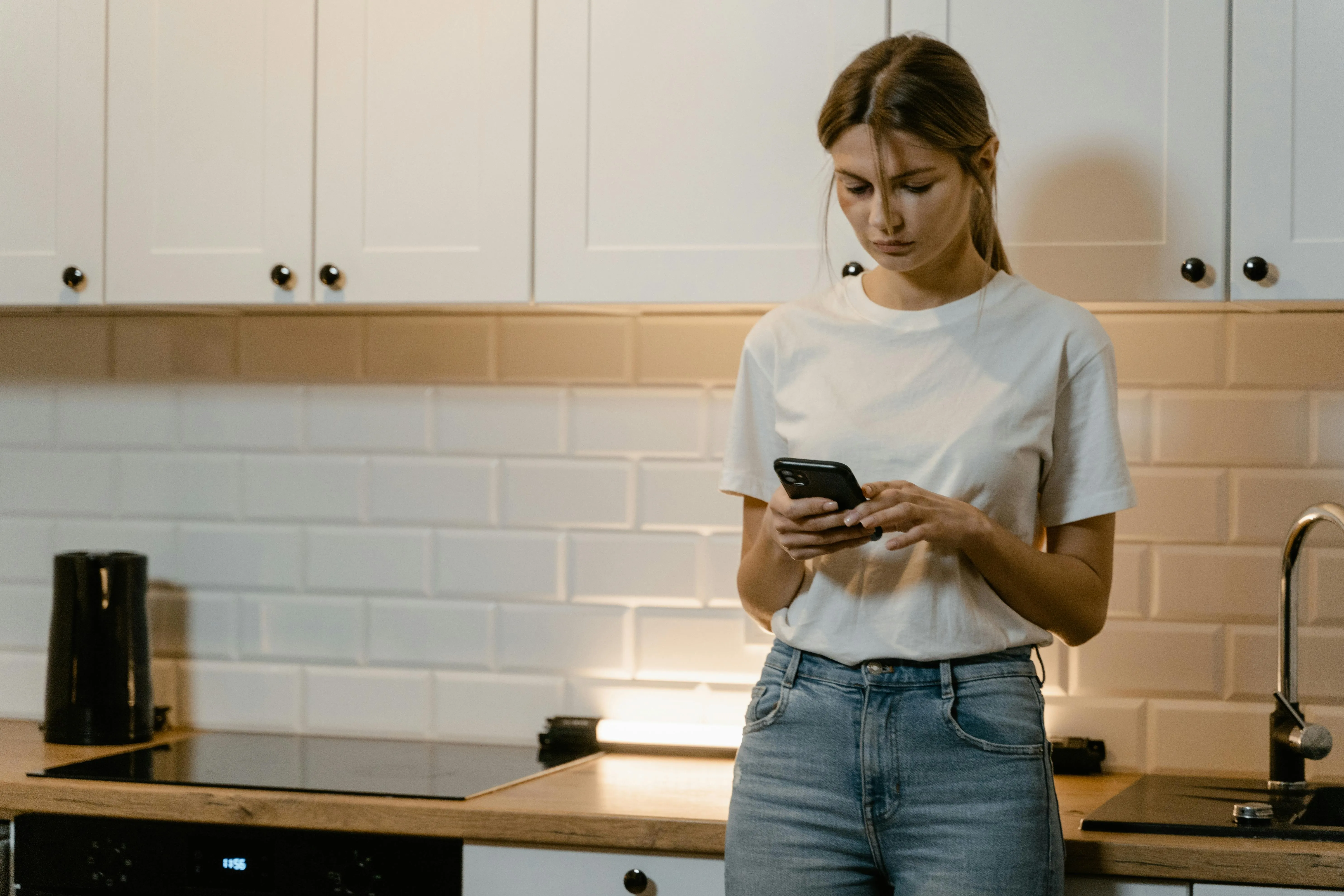 #FotoDescrição Mulher de camiseta clara e jeans, olhando para o celular com expressão séria, em uma cozinha moderna.