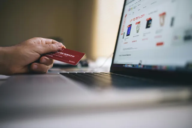 Mão segurando cartão de crédito em frente a laptop com site de compras.