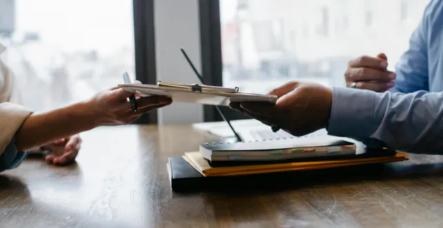 Duas pessoas trocando documentos sobre mesa com laptop e cadernos.