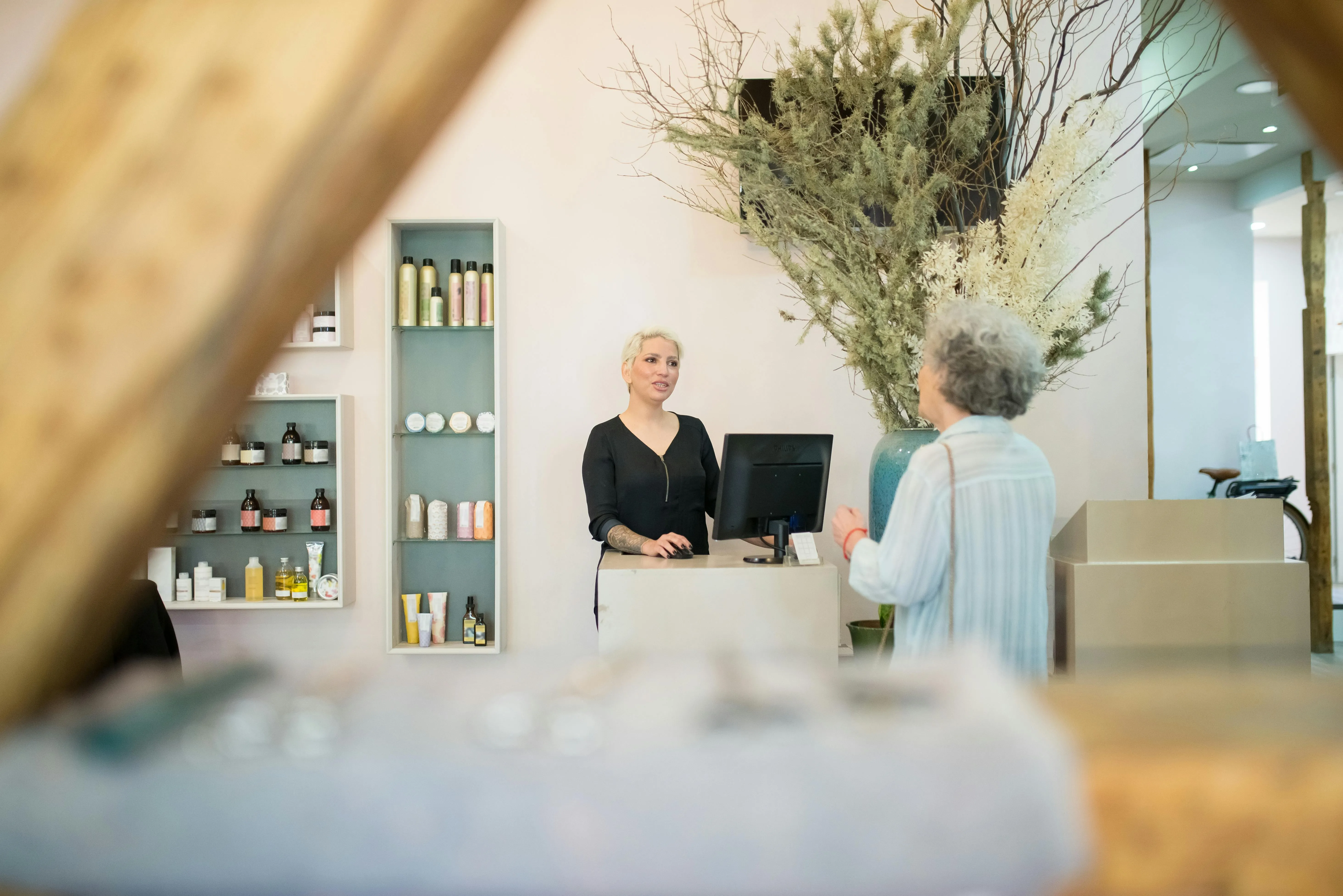 #FotoDescrição: Cliente sendo atendida por funcionária em recepção de salão de beleza moderno, com prateleira de produtos ao fundo.