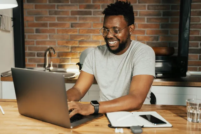 Pessoa sorrindo trabalhando no notebook em ambiente com parede de tijolos ao fundo