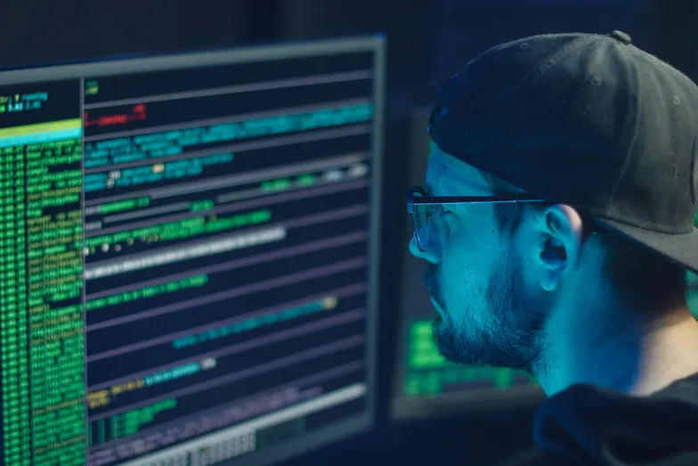 #FotoDescrição Homem com boné e óculos observando códigos em duas telas de computador em ambiente escuro.