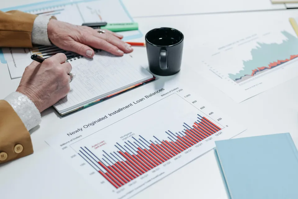 Pessoa anota dados financeiros ao lado de gráficos e xícara.