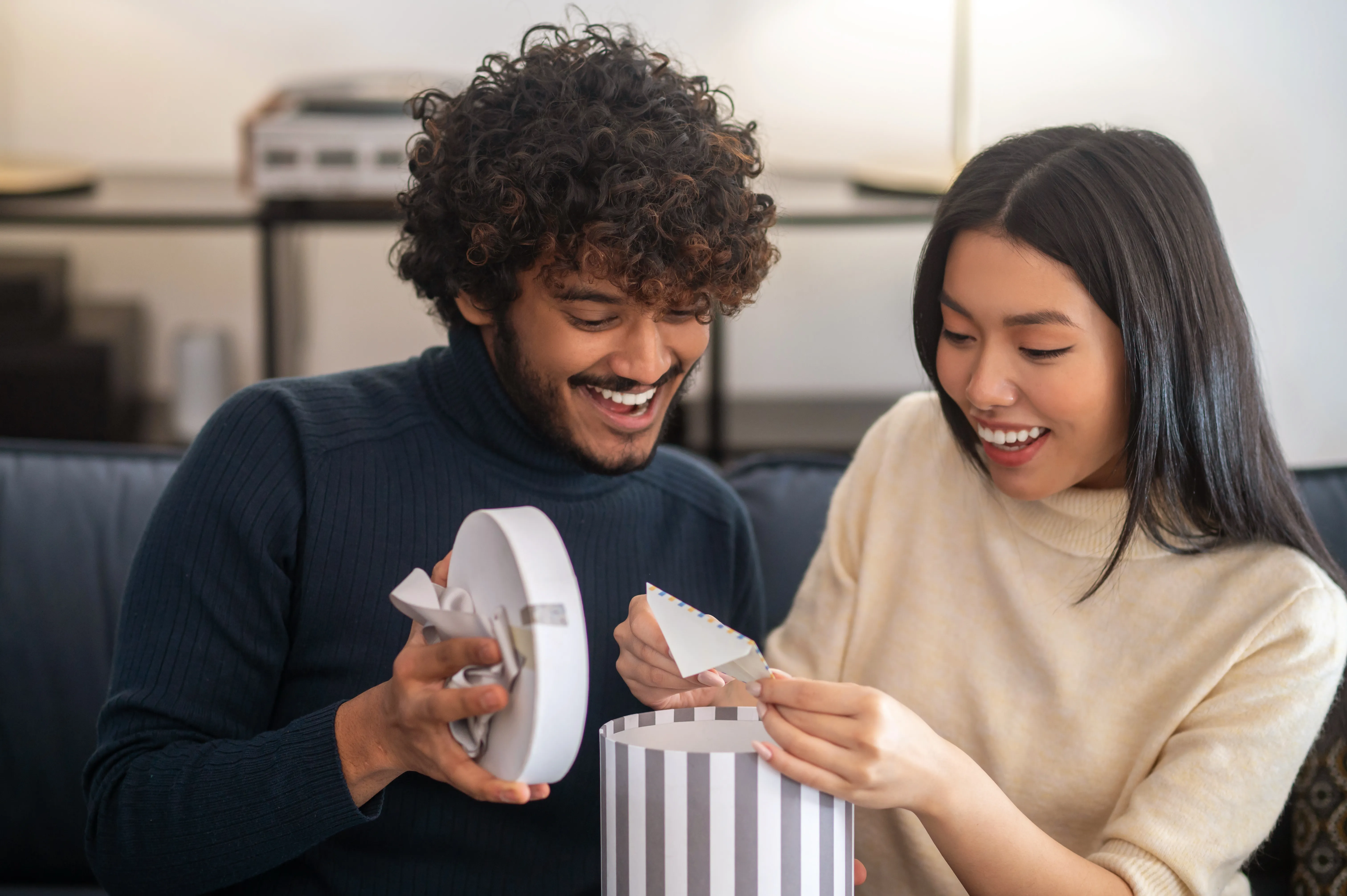 Casal jovem sorrindo no sofá enquanto abre um presente, iluminado por luz natural, criando um ambiente acolhedor.