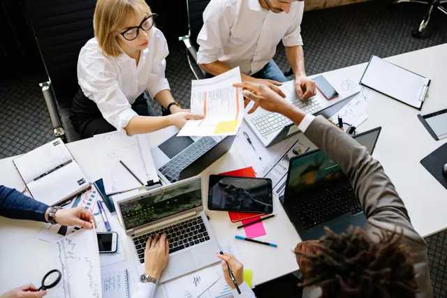 #FotoDescrição: Pessoas em uma mesa de trabalho colaborativo, compartilhando documentos e usando laptops e tablets.