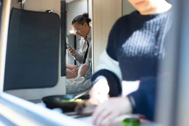 Pessoa usa celular dentro de van enquanto outra prepara comida na frente