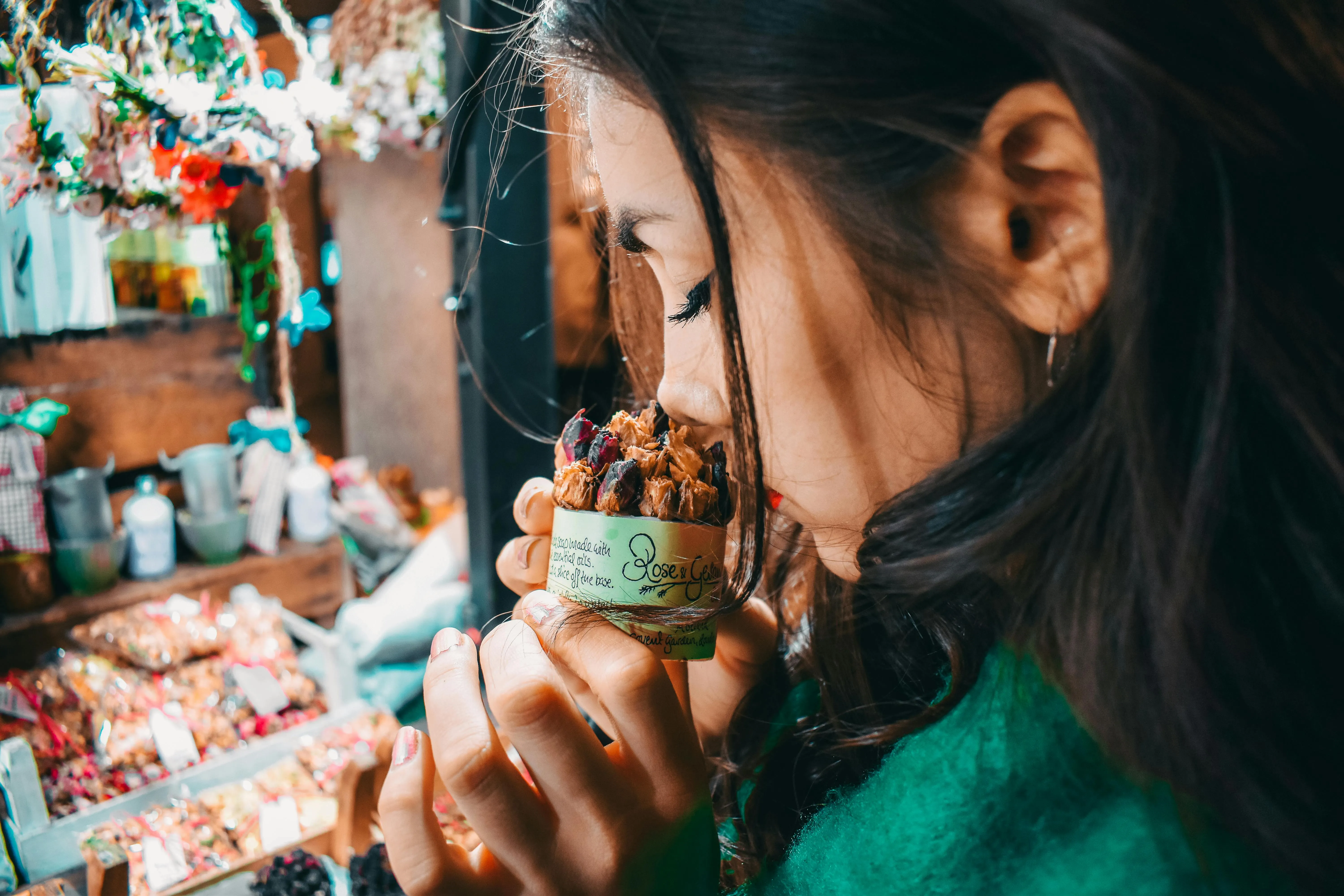 #FotoDescrição Mulher cheira um produto artesanal em um mercado local, com foco na experiência sensorial do olfato e no ambiente acolhedor e colorido do ponto de venda.