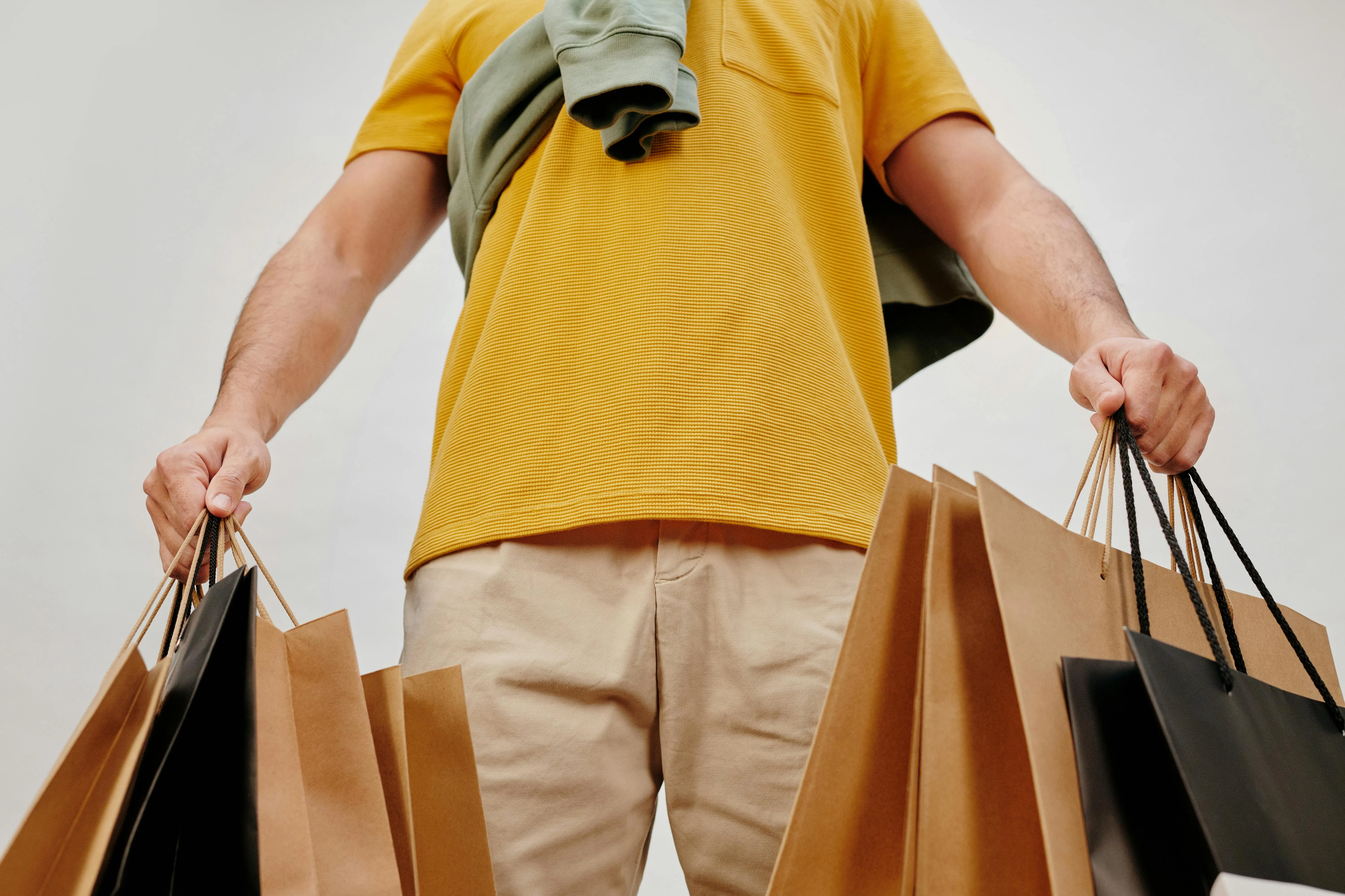 #FotoDescrição: Pessoa segurando várias sacolas de compras em ambos os braços, vestindo uma camiseta amarela e calça bege, contra um fundo branco.