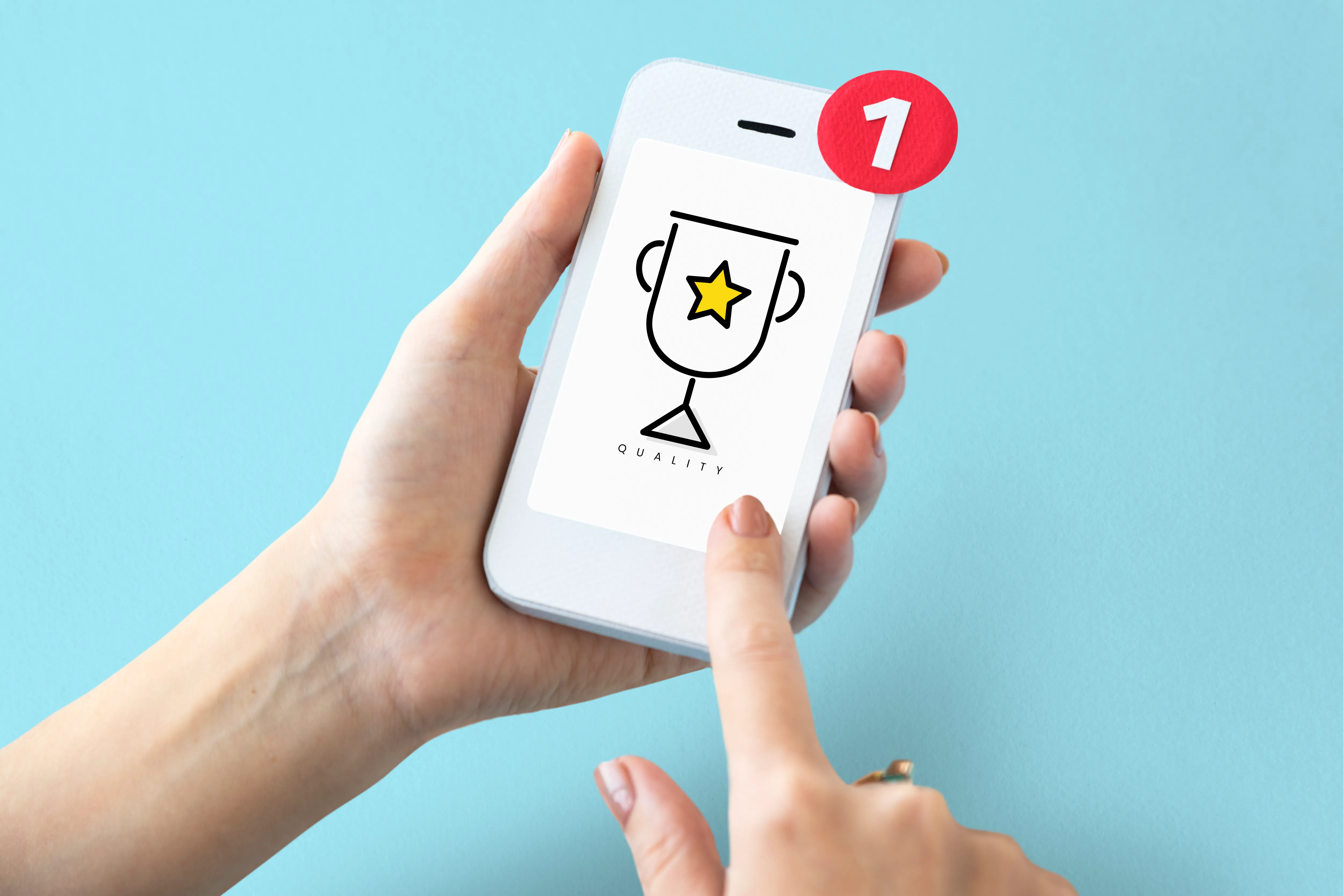 Uma mão segurando um smartphone exibe uma tela com um logotipo que apresenta um troféu com uma estrela amarela e a palavra "QUALITY" abaixo. Há uma notificação em vermelho no canto superior direito do celular, indicando uma nova mensagem ou alerta. O fundo da imagem é azul claro.