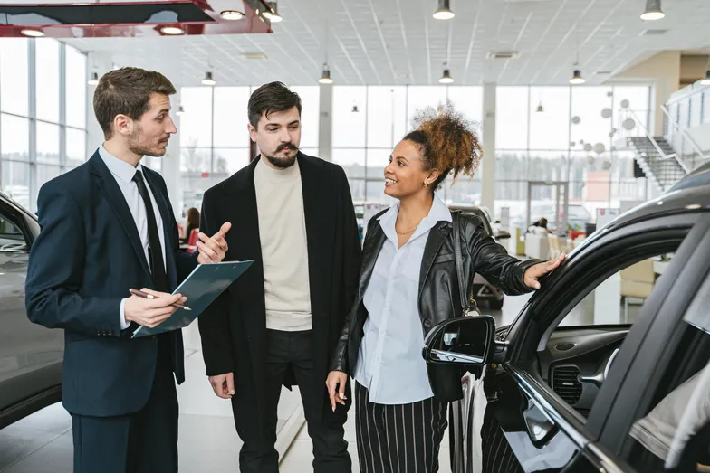 Três pessoas em uma concessionária discutindo sobre um carro, com um consultor de vendas apresentando informações.