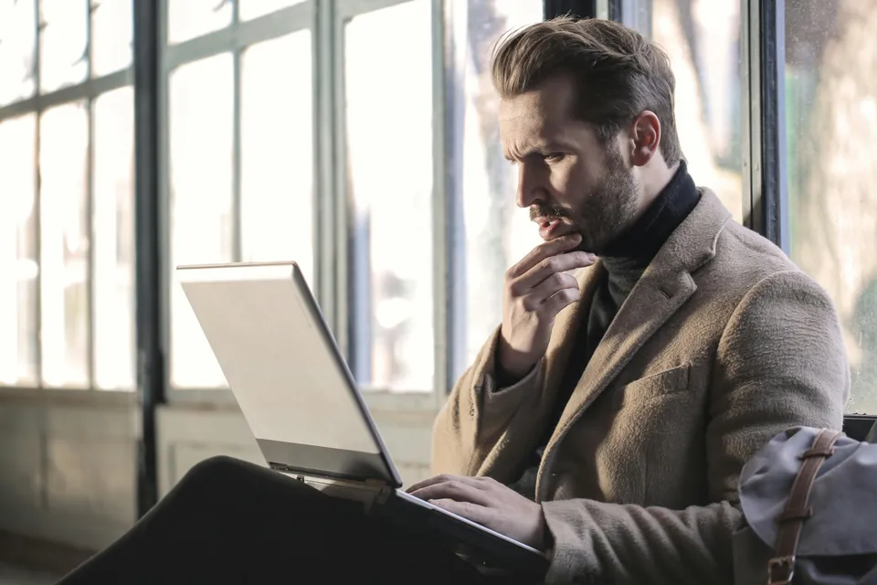 Homem pensativo digitando em notebook perto de grande janela clara.