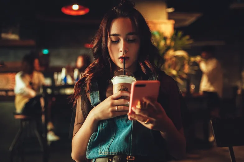 #FotoDescrição: Mulher jovem em um ambiente de cafeteria, segurando um copo de bebida com uma das mãos e um smartphone com a outra, enquanto está distraída navegando no telefone.