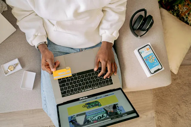 #FotoDescrição: Pessoa sentada em um sofá usando um notebook apoiado nas pernas, enquanto segura um cartão amarelo de crédito na outra mão. Ao lado, sobre o sofá, estão um celular com tela ligada, fones de ouvido e uma caixinha branca. A tela do notebook exibe uma promoção com destaque para "70% off".