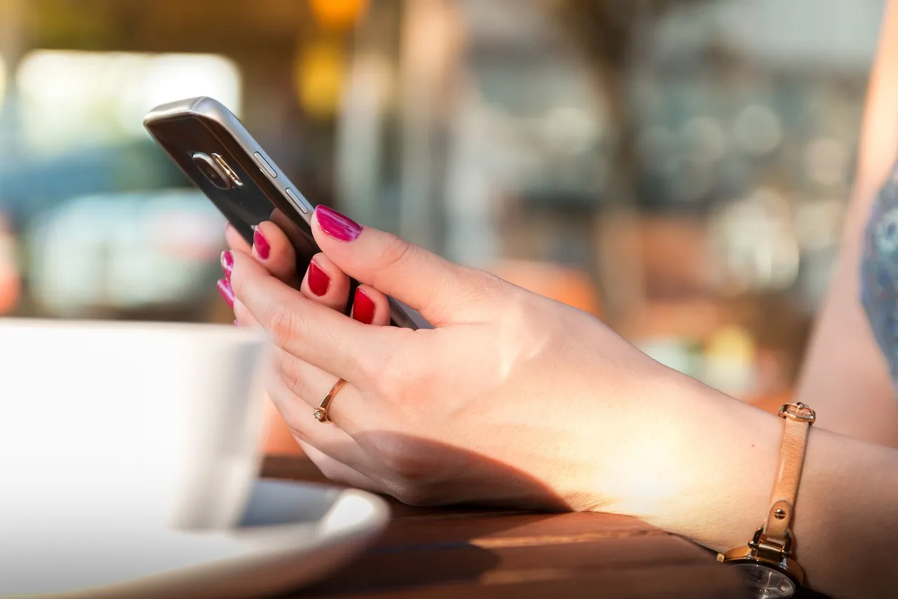 #FotoDescrição: Close-up de uma mão feminina segurando um smartphone em uma mesa de café. Representa interação digital e conveniência.