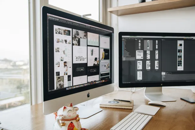 Mesa com dois monitores exibindo layouts de design gráfico.