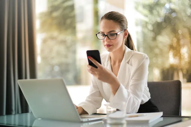 #FotoDescrição: Mulher de óculos em um escritório, analisando o celular enquanto trabalha no laptop, simbolizando produtividade e estratégia digital.