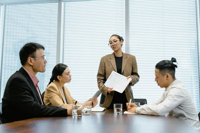 #FotoDescrição: Profissionais em reunião de negócios; uma mulher em pé apresenta um documento a três colegas atentos ao redor da mesa.