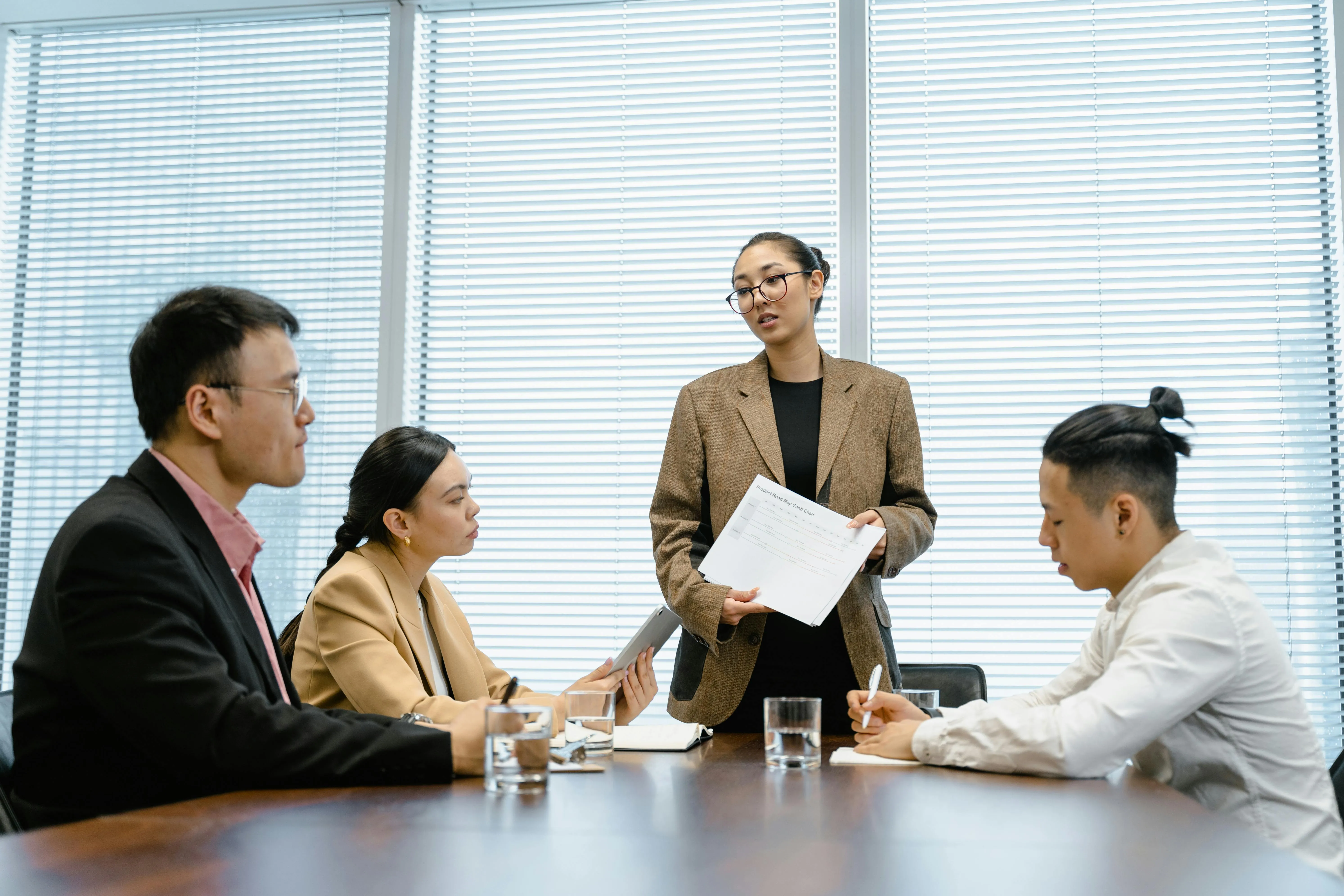 #FotoDescrição: Profissionais em reunião de negócios; uma mulher em pé apresenta um documento a três colegas atentos ao redor da mesa.