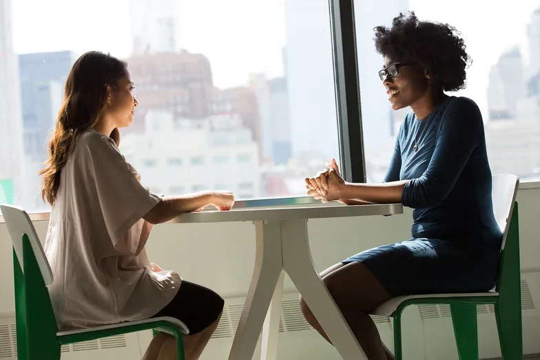 #FotoDescrição: Duas mulheres sentadas em uma mesa em frente a uma janela, conversando em um ambiente profissional.