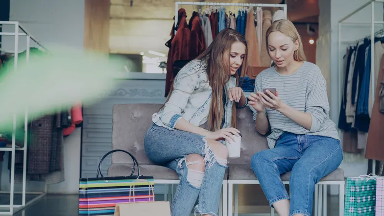Duas pessoas observam um celular em loja de roupas durante compras.
