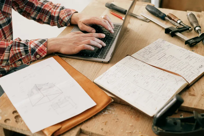 #FotoDescrição Pessoa digitando em um laptop sobre uma mesa com ferramentas e anotações.