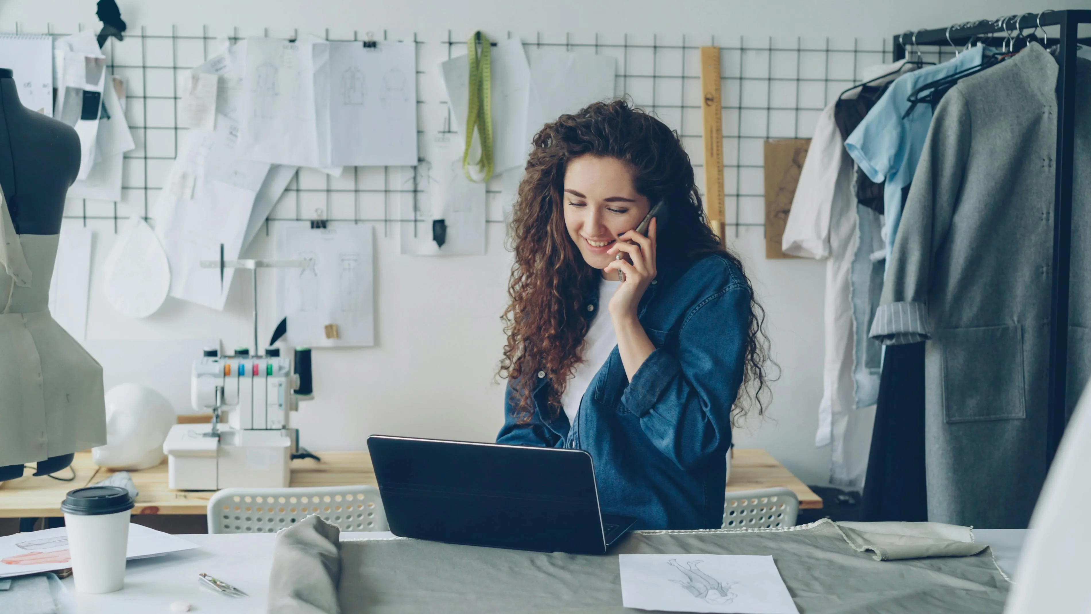 Designer de moda trabalha no laptop e fala ao telefone no ateliê