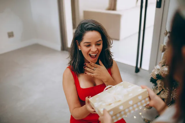 #FotoDescrição: Mulher surpresa e feliz recebendo um presente, simbolizando a satisfação.