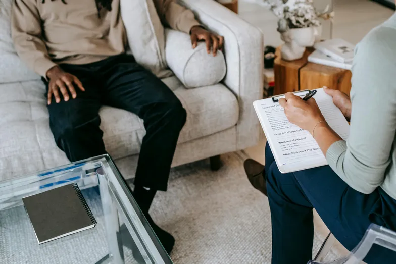 Sessão de terapia com duas pessoas, uma tomando notas e a outra sentada à frente, com uma mesa de vidro ao lado.