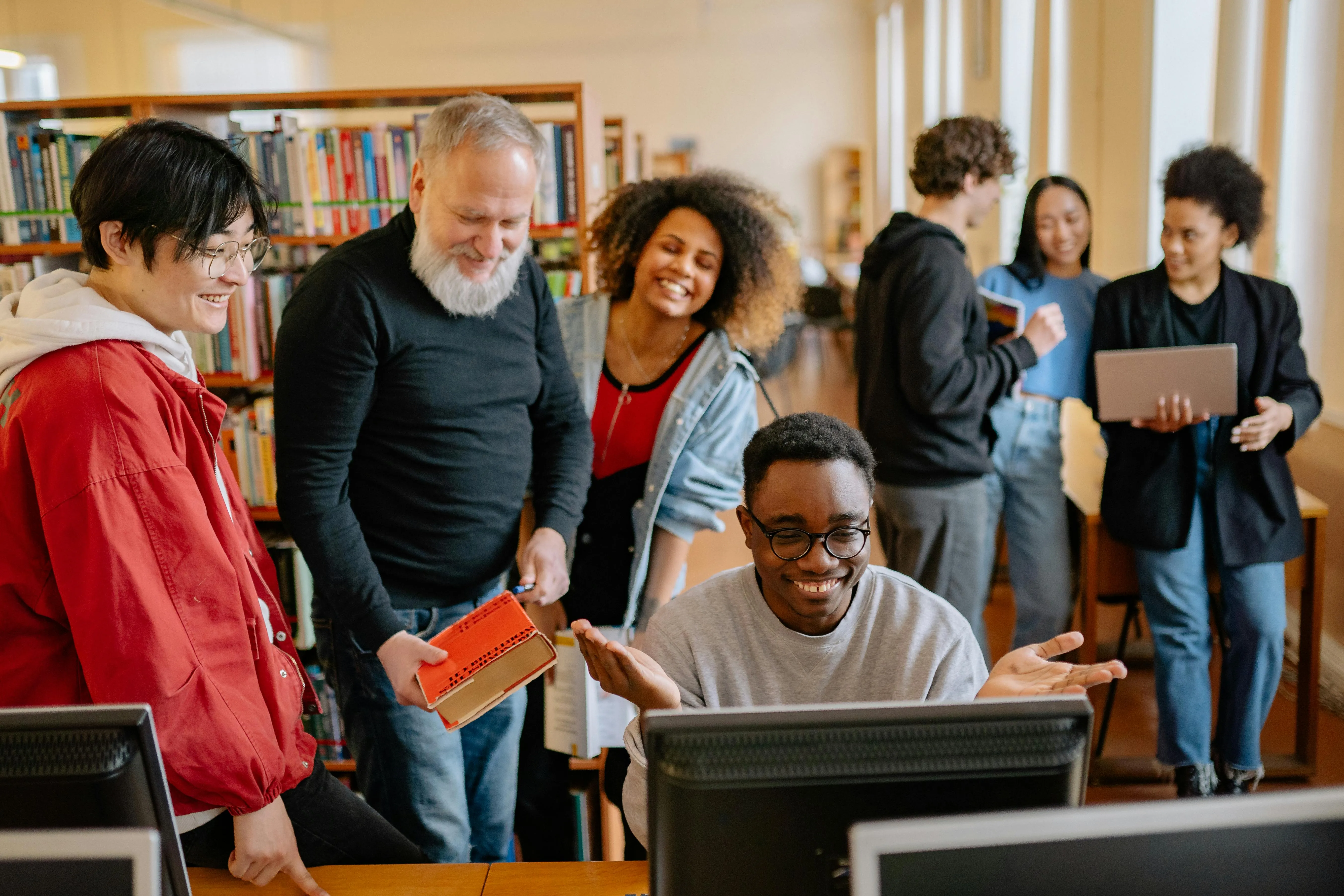 Pessoas em biblioteca rindo e conversando ao redor de um computador.