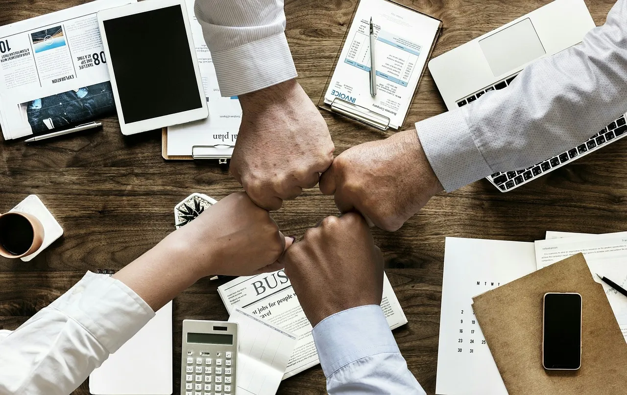 #FotoDescrição: Quatro pessoas unindo os punhos sobre uma mesa de trabalho, simbolizando trabalho em equipe, parceria e colaboração.