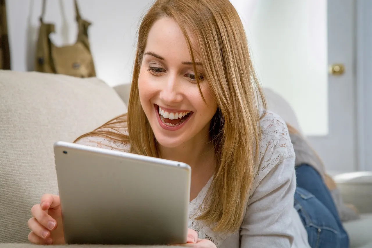 #FotoDescrição: Uma mulher sorrindo enquanto usa um tablet, destacando o uso de tecnologia digital e interação online.